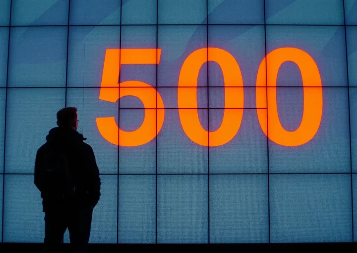 Eine Person steht vor einem großen digitalen Bildschirm, auf dem in fettem orangefarbenem Text die Zahl „500“ angezeigt wird, was wahrscheinlich auf einen HTTP 500-Fehler hinweist. Der Hintergrund besteht aus einem Raster aus beleuchteten blauen Quadraten, die einen auffälligen Kontrast erzeugen. Die Person scheint die Anzeige zu beobachten, die im Licht als Silhouette erkennbar ist.