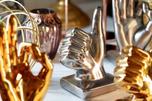 Eine Ausstellung verschiedener metallischer Handskulpturen auf einem Tisch. Die zentrale Skulptur ist eine polierte silberne Hand, die einen Daumen nach oben zeigt und damit echte Ergebnisse symbolisiert. Um sie herum sind goldene Handskulpturen in verschiedenen Posen und eine dekorative Vase im Hintergrund, die an die Eleganz der Web Awards erinnert.