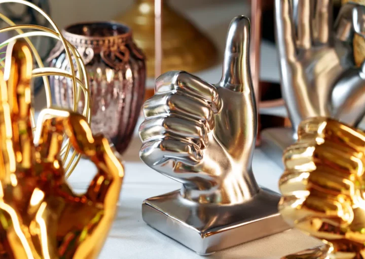 Eine Ausstellung verschiedener metallischer Handskulpturen auf einem Tisch. Die zentrale Skulptur ist eine polierte silberne Hand, die einen Daumen nach oben zeigt und damit echte Ergebnisse symbolisiert. Um sie herum sind goldene Handskulpturen in verschiedenen Posen und eine dekorative Vase im Hintergrund, die an die Eleganz der Web Awards erinnert.