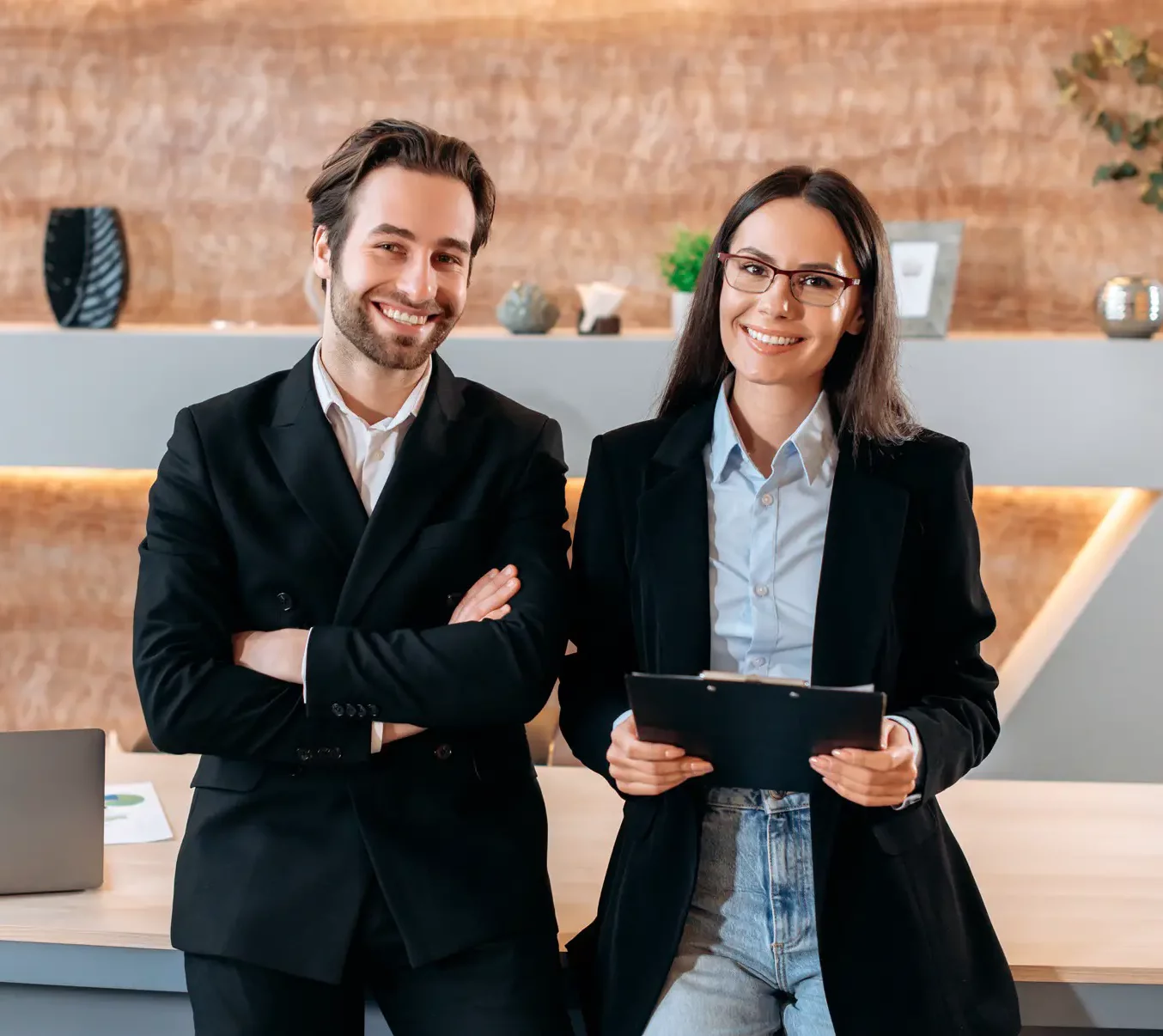 Zwei Berufstätige in Business-Kleidung stehen lächelnd in einem modernen B2B-Büro. Der Mann hat die Arme verschränkt, während die Frau ein Klemmbrett hält. Auf dem Holzschreibtisch vor ihnen liegt ein Laptop, was ihren kollaborativen Erfolgsansatz unterstreicht.
