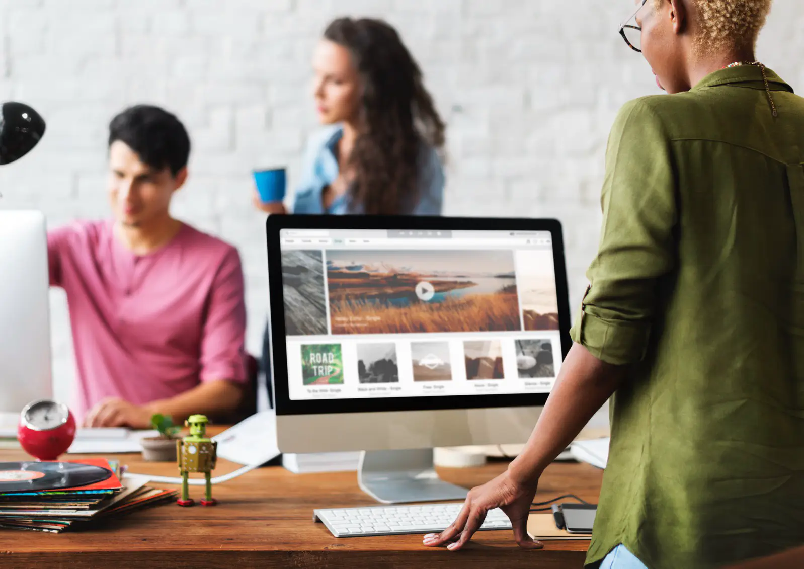 Eine Frau in einem grünen Hemd verwendet einen Desktop-Computer, auf dem Reisefotos angezeigt werden, möglicherweise für eine WordPress-Wartungsaufgabe. Im Hintergrund sitzt ein Mann in einem rosa Hemd neben einer Frau mit langen Haaren, die eine blaue Tasse hält. Auf dem Schreibtisch liegt ein kleines Roboterspielzeug, das der Szene Charme verleiht.