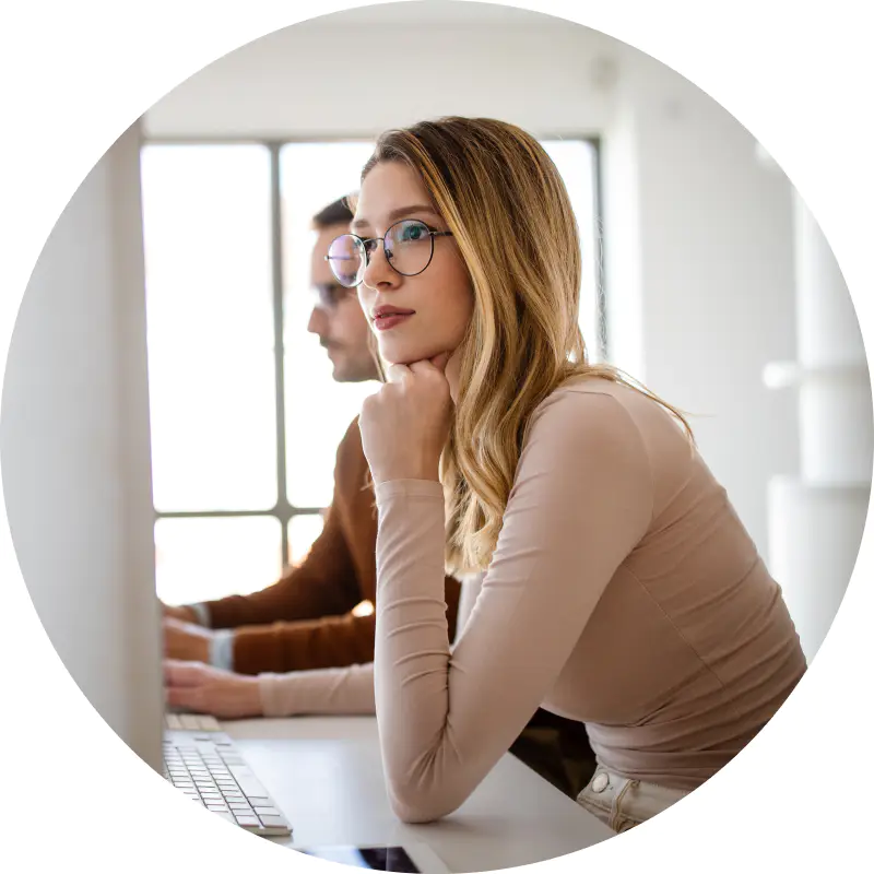 Eine Frau mit langen Haaren und Brille arbeitet konzentriert an einem Computer in einem hellen, modernen Büro und analysiert wahrscheinlich Marketingdaten. Eine weitere Person sitzt neben ihr und ist ebenso konzentriert auf ihren Bildschirm gerichtet. Die Szene lässt auf eine kollaborative oder konzentrierte Arbeitsumgebung schließen.