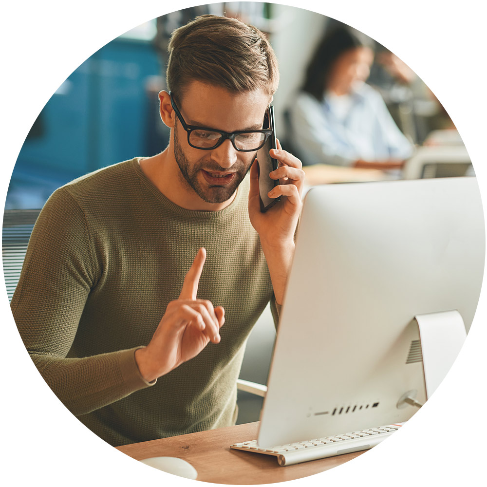 Ein Mann mit Brille sitzt an einem Schreibtisch und betreibt Telefonmarketing. Mit einer Hand telefoniert er auf seinem Smartphone, mit der anderen gestikuliert er, die Augen sind auf den Computermonitor gerichtet. Im Hintergrund ist eine verschwommene Büroumgebung zu sehen, in deren Nähe eine weitere Person sitzt.
