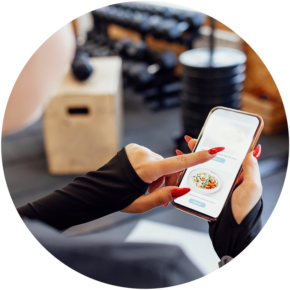 Im Fitnessstudio wählt eine Person mithilfe einer Smartphone-App wie einer digitalen Microsite ihre nächste Mahlzeit aus. Auf dem Bildschirm wird eine Schnittstelle zur Essensplanung angezeigt, während im Hintergrund Fitnessgeräte wie Gewichte und eine Holzkiste zu sehen sind.