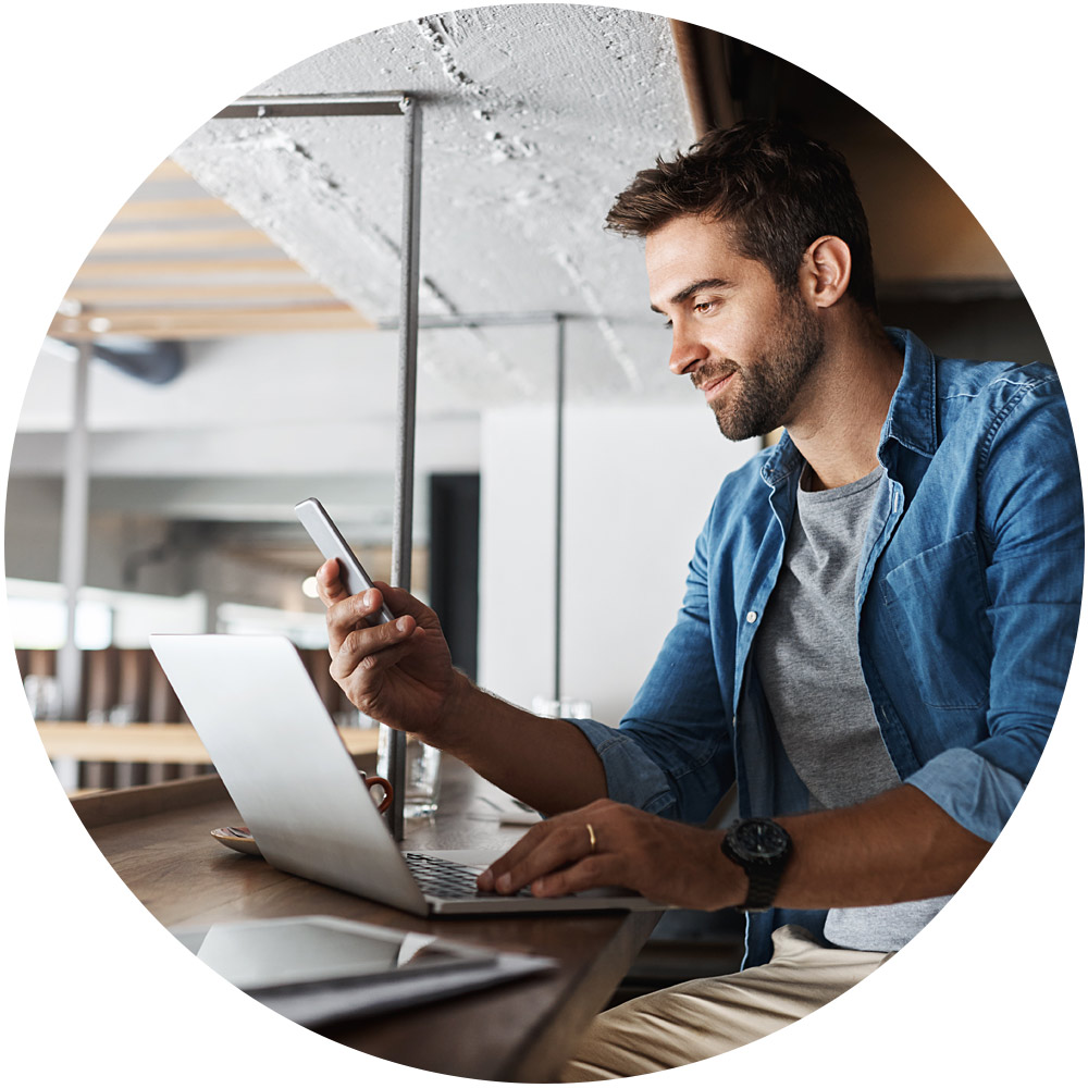Ein Mann in legerer Kleidung sitzt an einem Tisch in einem modernen Café, benutzt seinen Laptop und hält ein Smartphone in der Hand. Er lächelt begeistert, während er sich in die App-Erstellung vertieft. Das helle Ambiente wird von natürlichem Licht erhellt, das durch große Fenster hereinfällt.