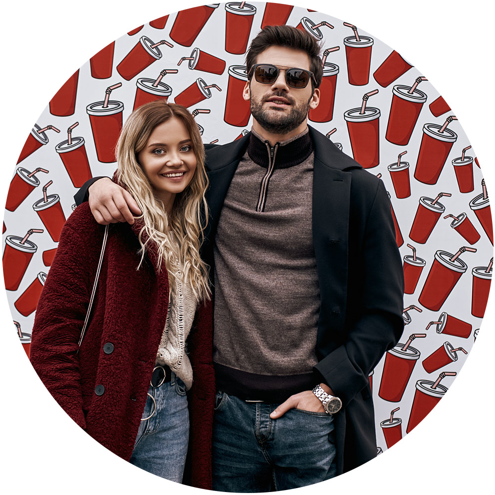Ein lächelndes Paar steht vor einer Wand, die mit einem Corporate Design mit roten Getränkebechern geschmückt ist. Die Frau mit den langen blonden Haaren trägt einen burgunderfarbenen Mantel, während der Mann mit kurzen Haaren und Sonnenbrille einen schwarzen Mantel trägt und mit seinem Arm um ihre Schulter posiert.