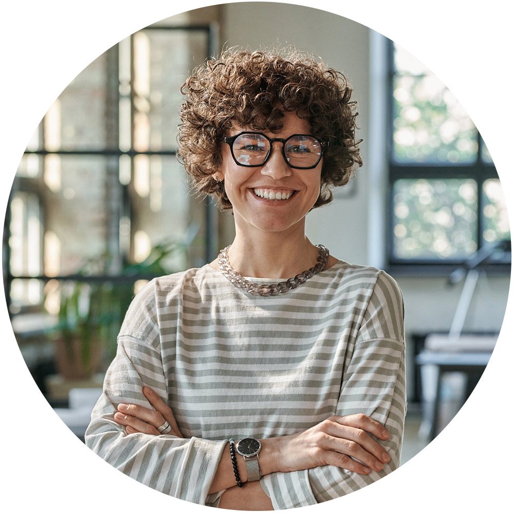 Eine Person mit lockigem Haar und Brille, die die Essenz von Marketing-Funnels verkörpert, steht lächelnd mit verschränkten Armen in einem hellen Raum. Sie trägt ein gestreiftes Hemd und Armbänder, ergänzt durch große Fenster und im Hintergrund sind grüne Pflanzen zu sehen.