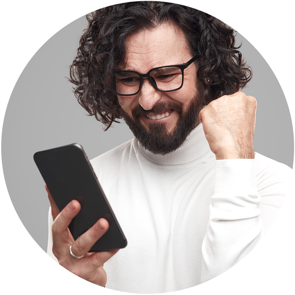 Ein bärtiger Mann mit lockigem Haar und Brille blickt mit freudigem Gesichtsausdruck auf ein Smartphone und schwelgt möglicherweise in den Erfolgen der Gamifizierung. Er trägt einen weißen Rollkragenpullover und streckt feierlich seine Faust in die Höhe, umrahmt von einem schlichten grauen Kreis.