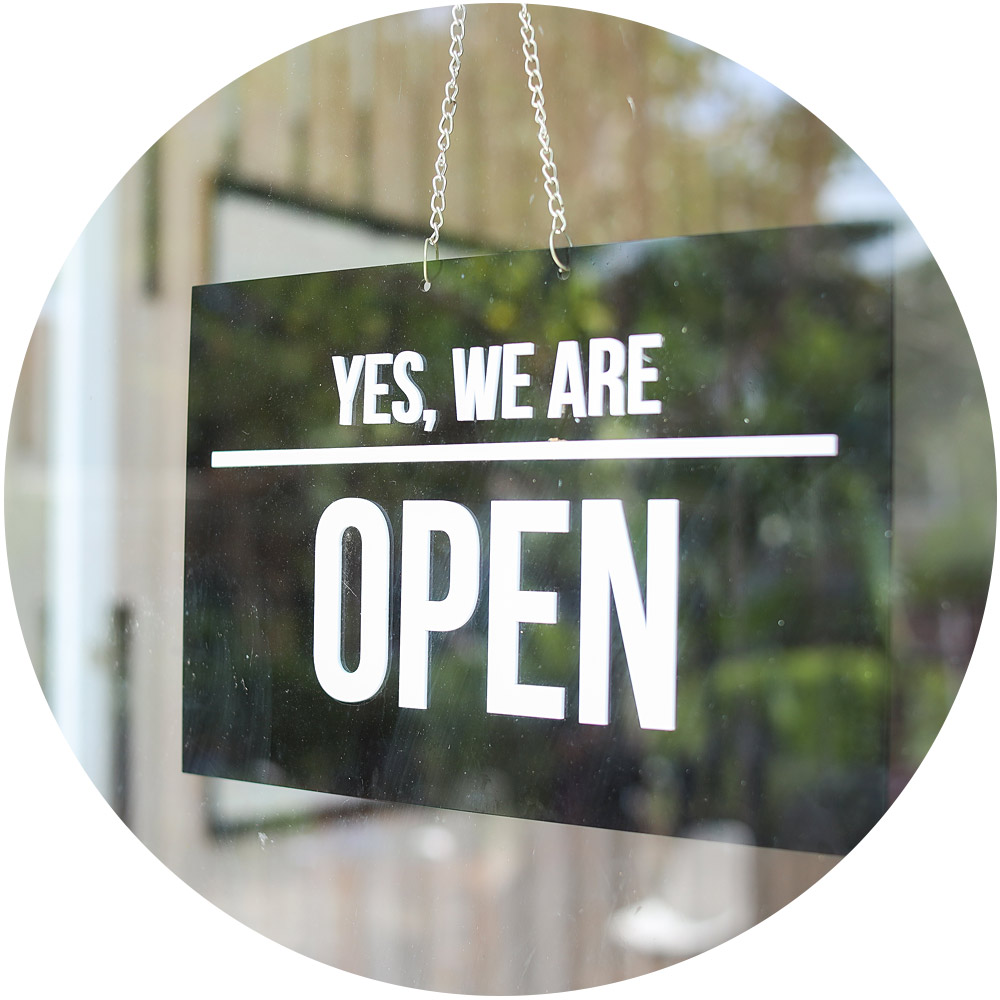 Ein schwarzes Schild mit weißem Text verkündet stolz „Ja, wir haben geöffnet“ im Fenster und lädt Besucher ein, die Dienste zu erkunden, die SEO- und Smart Lead-Strategien optimieren. Dahinter sorgt eine verschwommene Außenszene mit viel Grün für eine erfrischende Note.