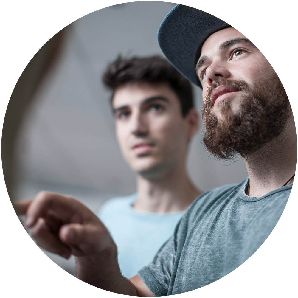 Zwei junge Männer diskutieren miteinander. Einer trägt eine Mütze und zeigt auf etwas außerhalb des Bildes. Das Bild konzentriert sich auf ihre Ausdrücke und vermittelt Konzentration und Zusammenarbeit bei intelligenten Führungsstrategien in einem kreisförmigen Rahmen.
