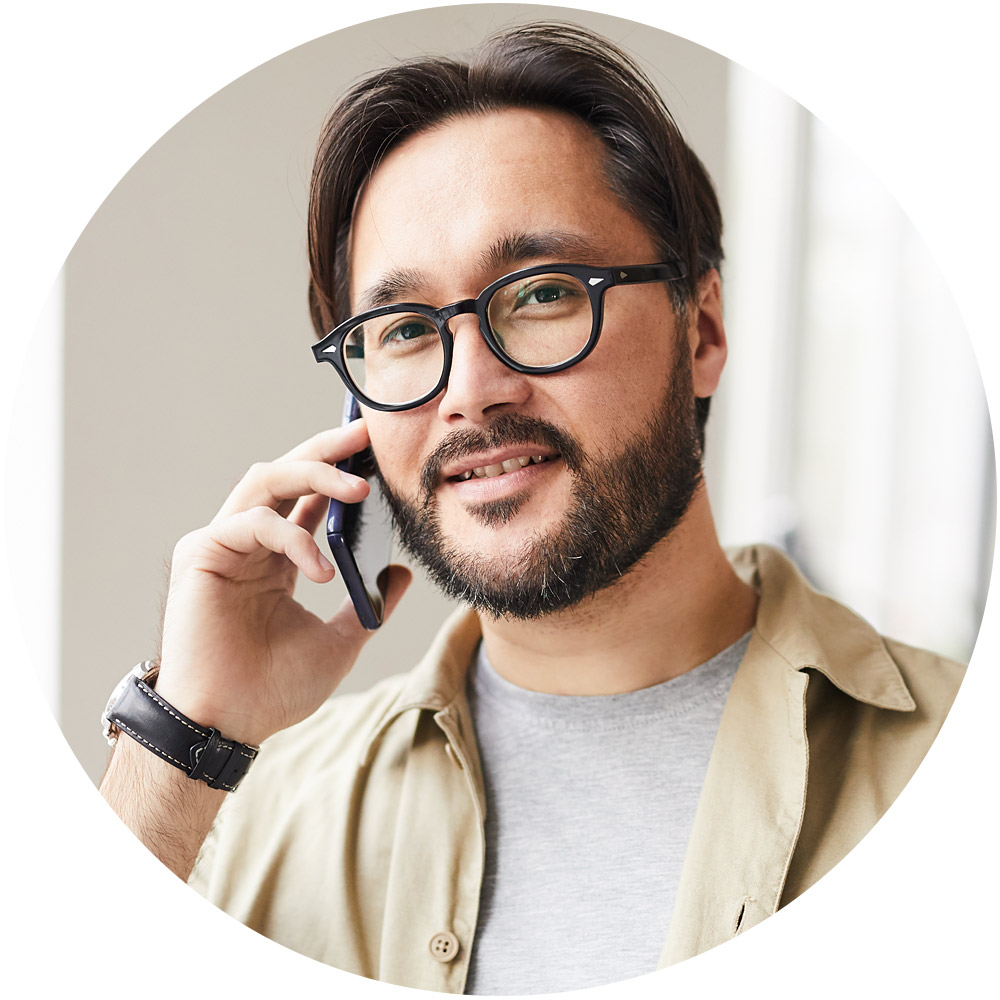 Ein Mann mit Brille und Bart lächelt, während er über sein Smartphone über einen Dienst spricht. Er trägt ein beiges Hemd über einem grauen T-Shirt und steht in einer Innenumgebung mit leicht verschwommenem Hintergrund, möglicherweise in der Nähe des Fensters neben dem Briefkasten.