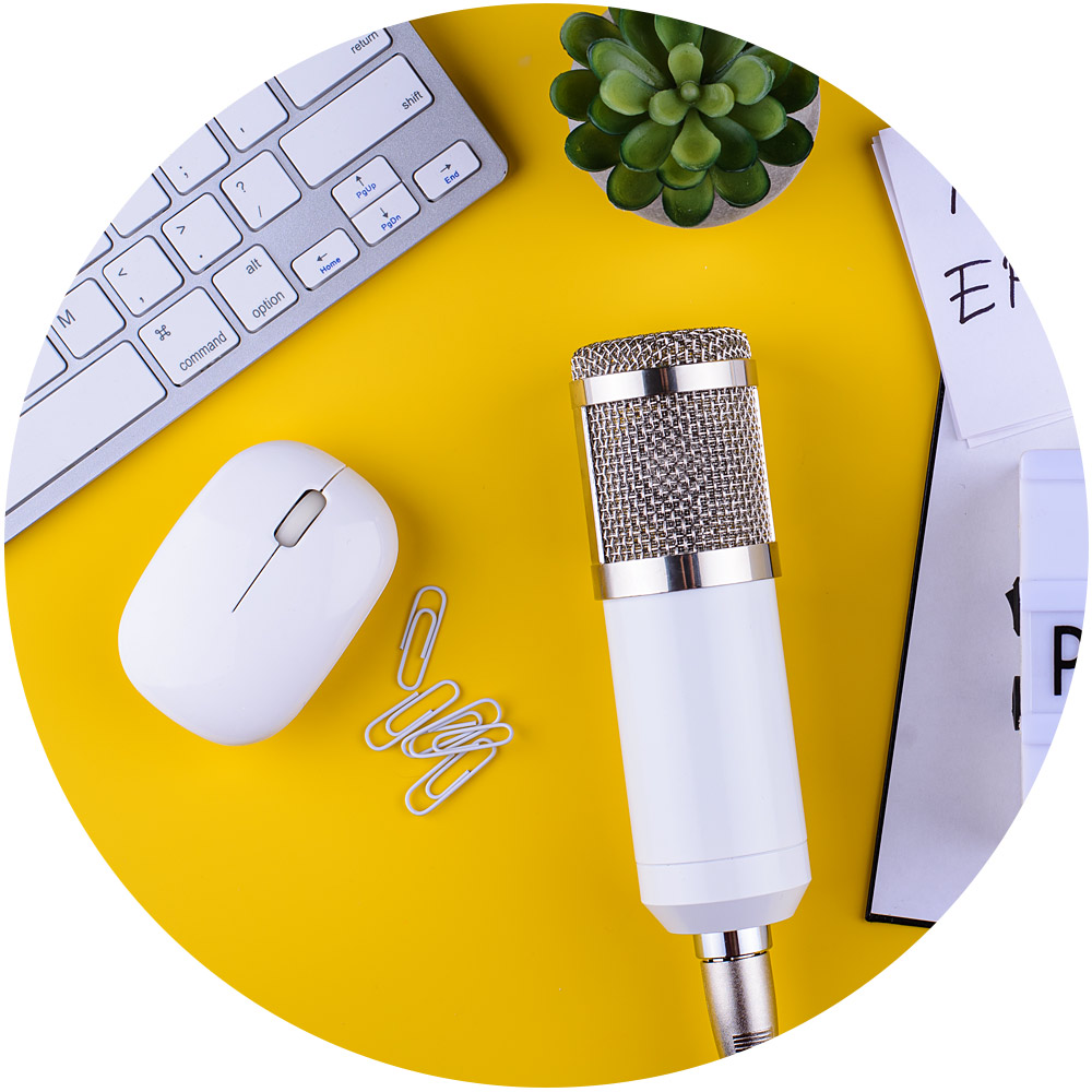 Ein weißes Mikrofon auf einer leuchtend gelben Oberfläche steht umgeben von einer Computertastatur, einer Maus, Büroklammern und verstreuten Blättern Papier. Eine kleine Topfpflanze verleiht diesem lebendigen Setup einen Hauch von Grün und ist perfekt, um die Essenz Ihrer Markenstimme in jeder Aufnahme einzufangen.