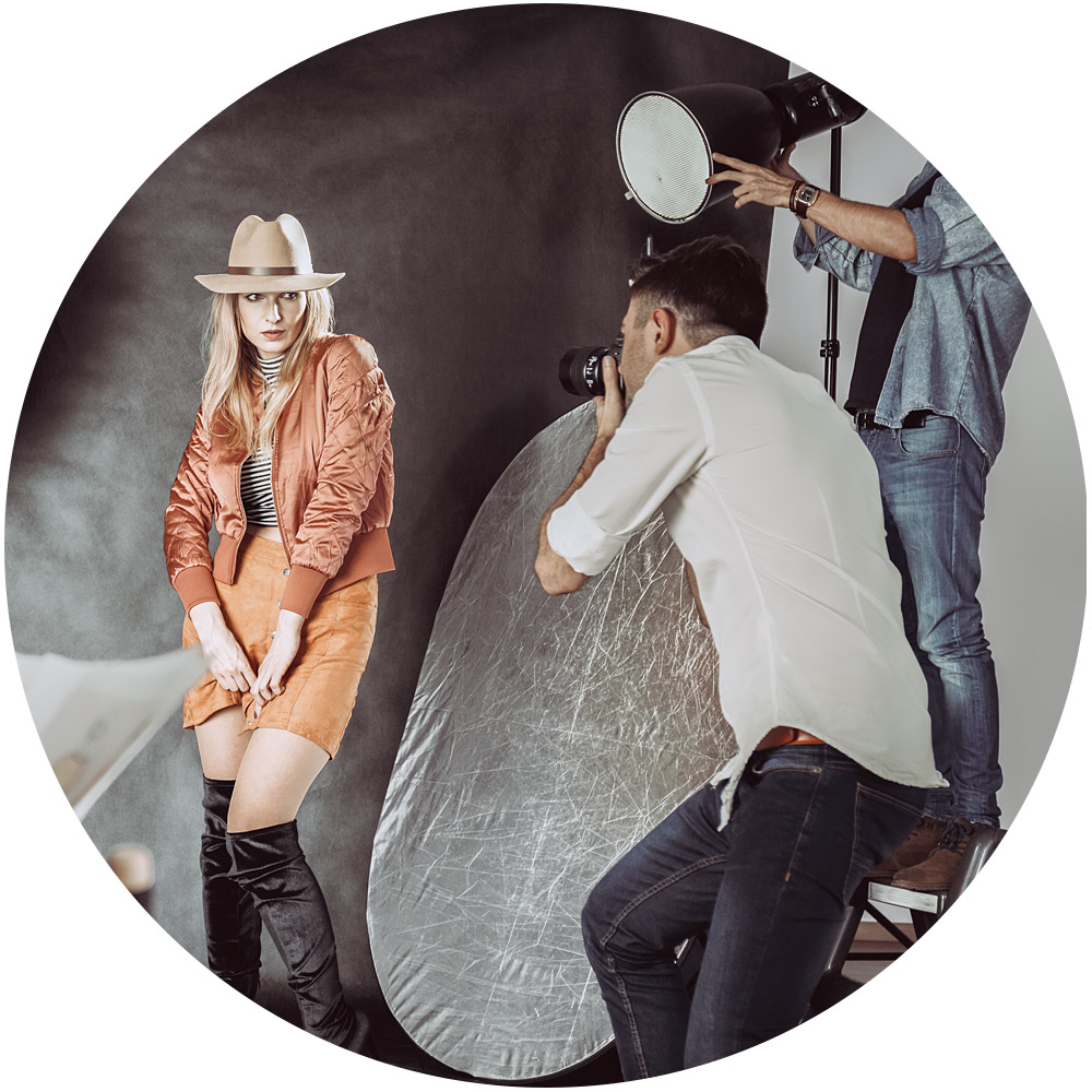 Ein Model posiert elegant in einem Studio. Es trägt einen beigen Hut, eine orangefarbene Jacke, einen braunen Rock und schwarze kniehohe Stiefel. Während des Shootings nimmt ein Fotograf ihr Bild auf, während ein Assistent die Beleuchtung einstellt und dabei eine große reflektierende Platte hält.