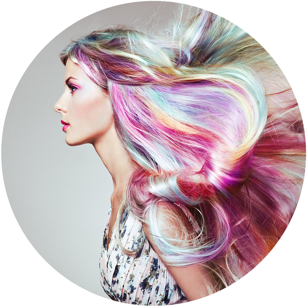 Ein Model mit langen, wallenden Haaren in leuchtenden Regenbogenfarben, darunter Pink, Blau, Lila und Gelb. Sie tragen ein gemustertes Oberteil und leuchtendes Make-up und stehen vor einem schlichten Hintergrund. Das Bild ist kreisförmig.