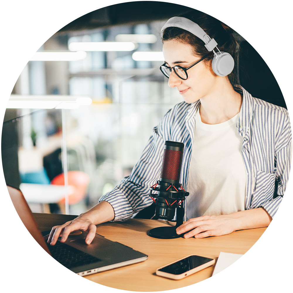 Eine Person mit Kopfhörern arbeitet an einem Laptop an einem für Podcasts ausgestatteten Schreibtisch mit Mikrofon und Smartphone. Die moderne Büroumgebung strahlt Professionalität aus, wobei die unscharfe Hintergrundbeleuchtung zur Atmosphäre beiträgt.