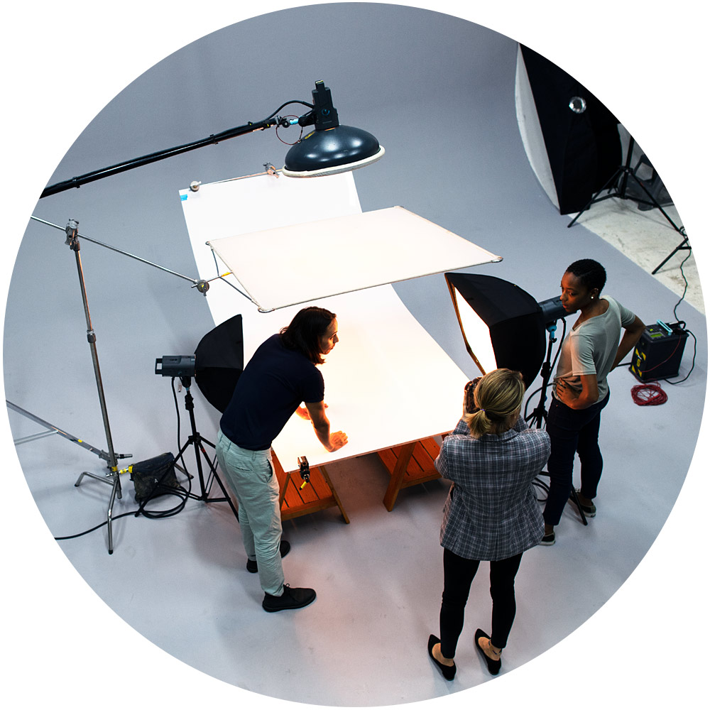 Drei Personen bereiten ein Fotoshooting für Produktfotos in einem Studio vor. Einer richtet den Hintergrund auf einem Tisch aus, während die anderen zuschauen. Um sie herum sind Studiolichter und Requisiten sichtbar, was eine professionelle und kollaborative Atmosphäre schafft, die perfekt für die Aufnahme eindrucksvoller Produktbilder ist.