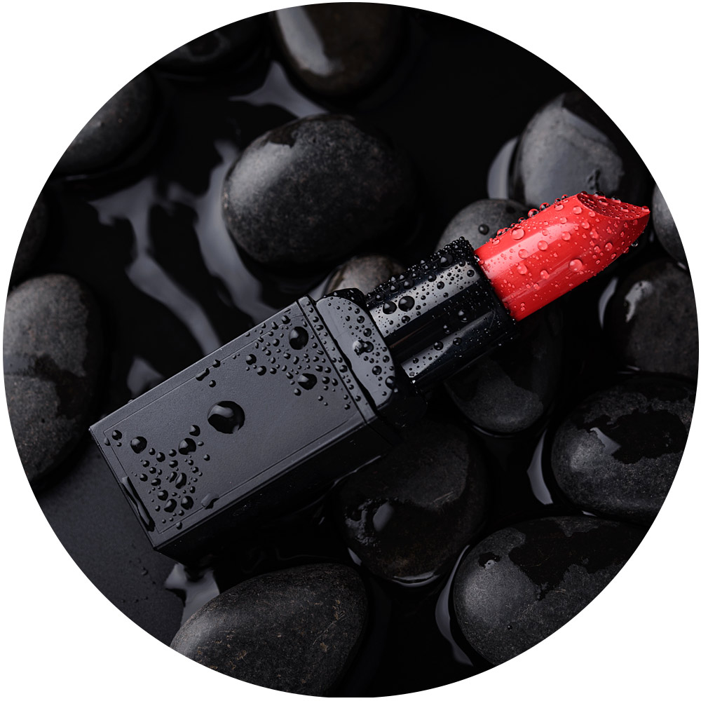 Ein schwarzer Lippenstift mit einer leuchtend roten Spitze glitzert unter Wassertropfen und erzeugt atemberaubende Produktfotos, wenn er auf glatten, nassen schwarzen Steinen ruht. Das glänzende Rot hebt sich deutlich von den dunklen Steinen ab und erzeugt einen auffälligen Kontrast.