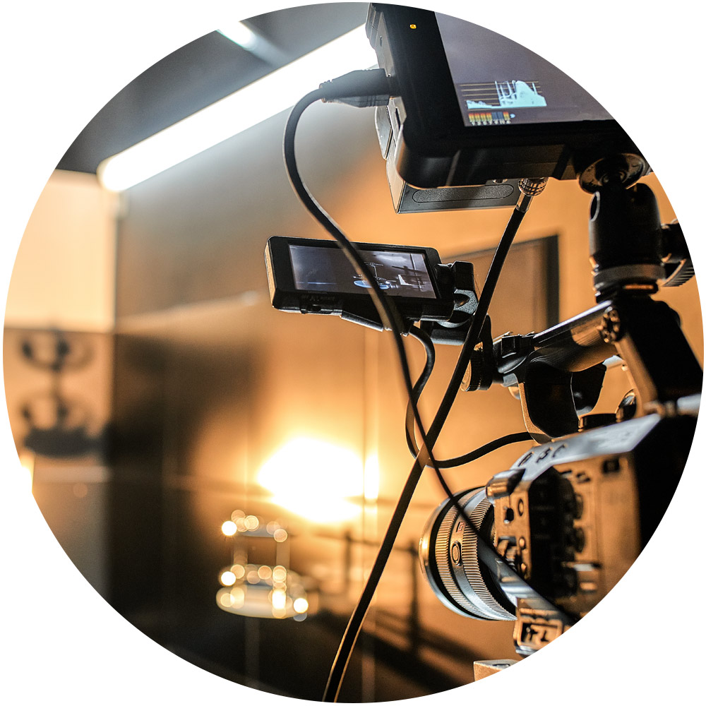 Nahaufnahme einer professionellen Kamera auf einem Stativ in einem schwach beleuchteten Studio. Die Kamera ist über Kabel mit mehreren Monitoren verbunden und fängt eine warme, beleuchtete Szene im Hintergrund ein – perfekt für die Erstellung überzeugender Produktvideos.