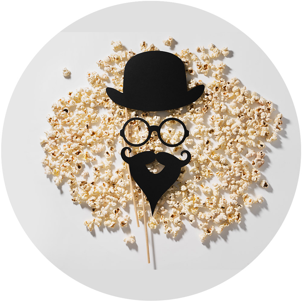 Schwarze Ausschnitte eines Hutes, einer runden Brille und eines Schnurrbarts auf Stöcken sind auf einem verstreuten Hintergrund aus Popcorn angeordnet und imitieren ein Gesicht. Die Gegenstände werfen subtile Schatten und verleihen dem verspielten Design Tiefe, das sich perfekt für Produktvideos eignet.