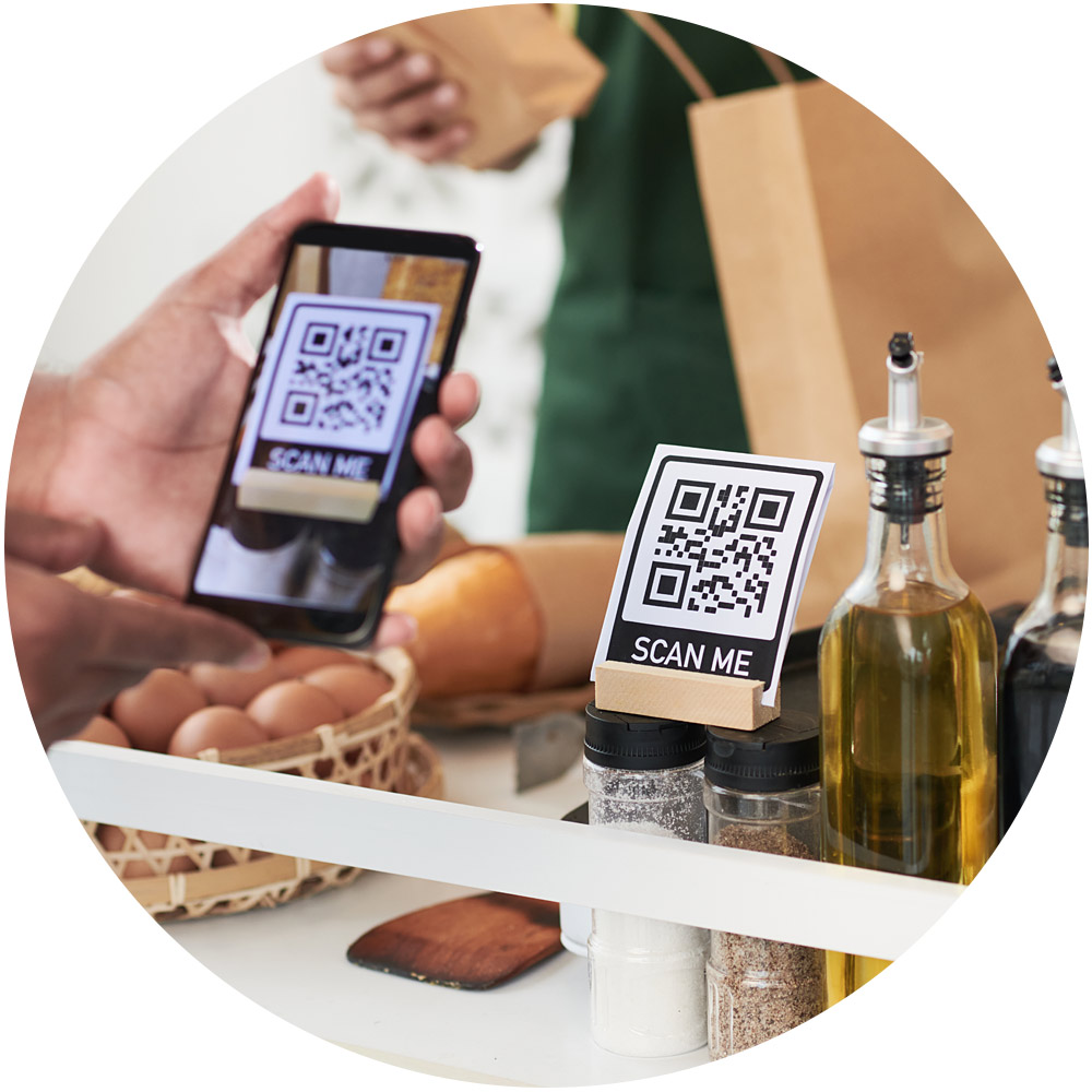 Eine Person scannt mit einem Smartphone an einer Ladentheke einen dynamischen QR-Code mit der Aufschrift „Scan Me“. In der Nähe sind verschiedene Artikel zu sehen, darunter ein Korb mit Eiern, eine braune Papiertüte sowie Flaschen mit Speiseöl und Gewürzen.