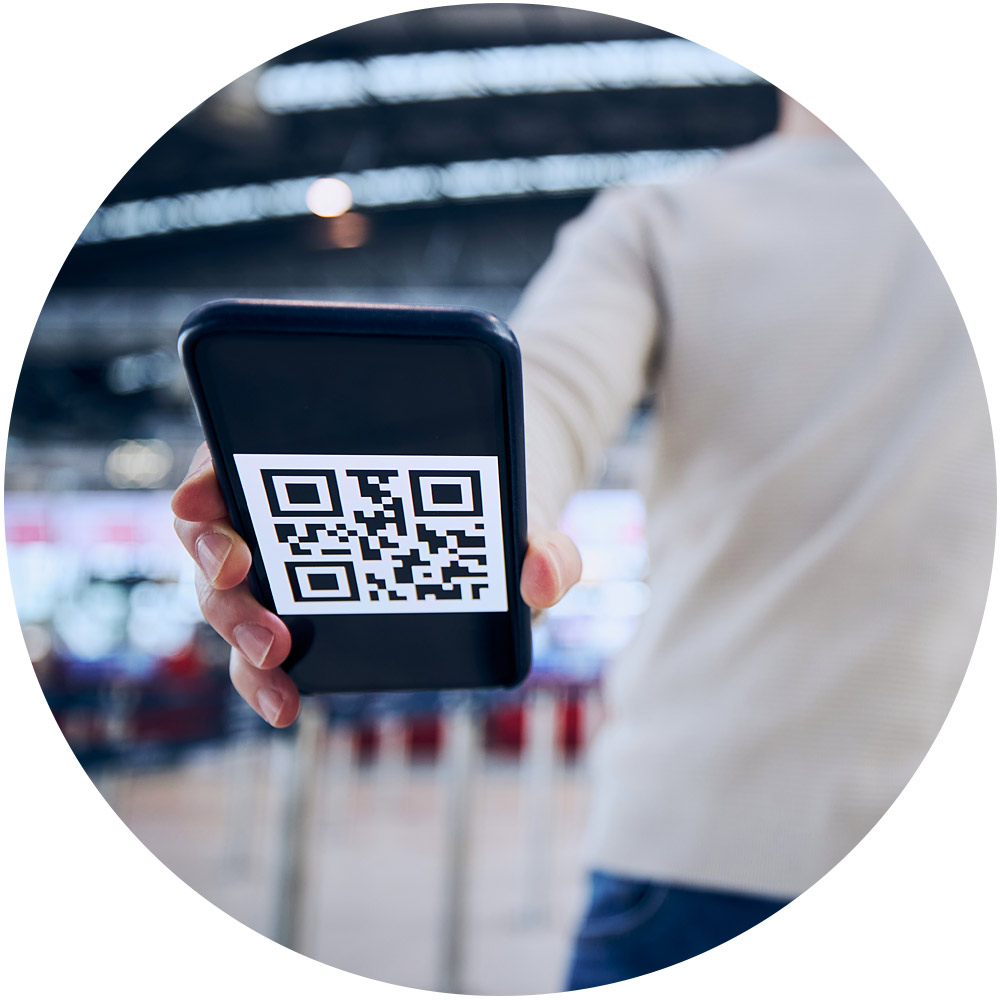 Eine Person hält ein Smartphone, auf dem ein dynamischer QR-Code angezeigt wird. Der Hintergrund ist ein verschwommener öffentlicher Ort, möglicherweise ein Flughafen oder Bahnhof, mit hellen Lichtern und Strukturen in der Ferne.