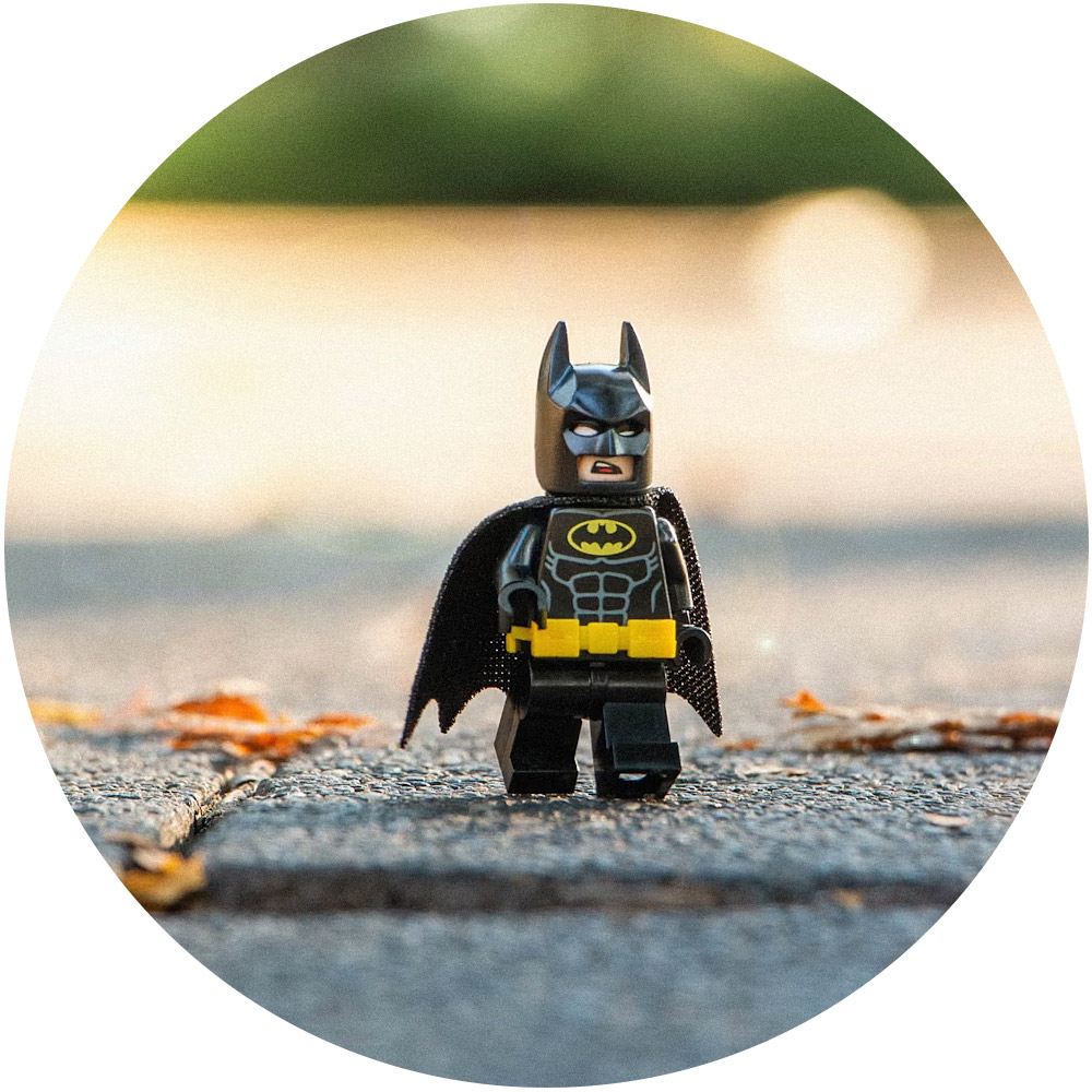 Eine LEGO Batman-Figur steht auf einer gepflasterten Fläche, auf der Herbstblätter verstreut sind. Der Hintergrund ist unscharf, wodurch ein Bokeh-Effekt entsteht, der den Ruf dieses Mini-Superhelden gekonnt vermittelt und seinen schwarzen Anzug mit dem ikonischen gelben Fledermaussymbol hervorhebt.