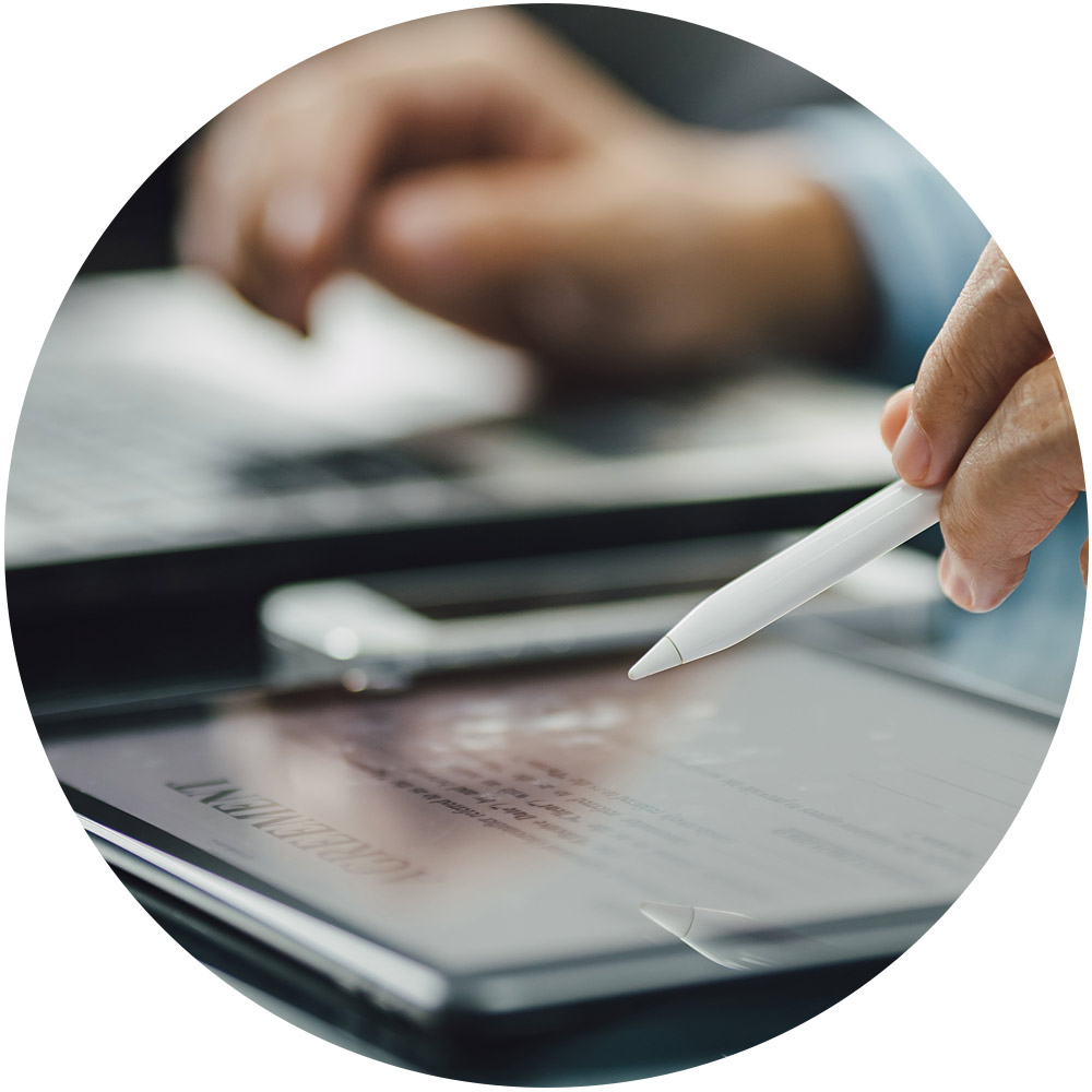 Eine Hand mit einem Stift schwebt über einem Tablet-Bildschirm, auf dem Text angezeigt wird, und deutet damit auf einen Signaturdienst in Aktion hin. Im Hintergrund steht ein Laptop auf einem Schreibtisch und vervollständigt die Szene eines modernen, digitalen Arbeitsplatzes.