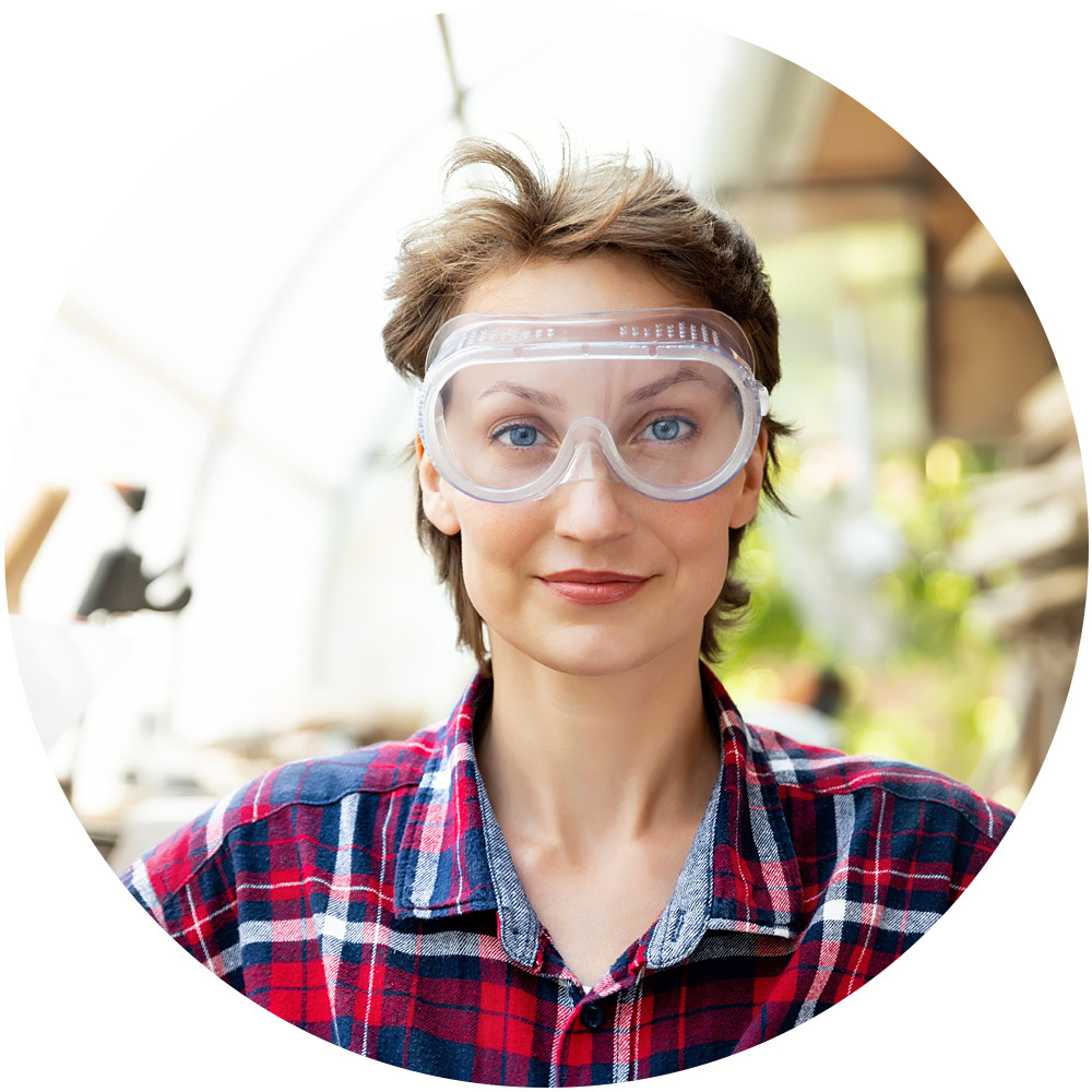 Eine Person mit einer durchsichtigen Schutzbrille und einem rotkarierten Hemd steht drinnen und strahlt das Selbstvertrauen und die Konzentration aus, die für jeden Einstellungsprozess ideal sind. Der sanft verschwommene Hintergrund lässt an ein Gewächshaus oder eine Industrieumgebung denken und eignet sich perfekt, um ihre Bereitschaft in einer Einstellungsumgebung zu demonstrieren.