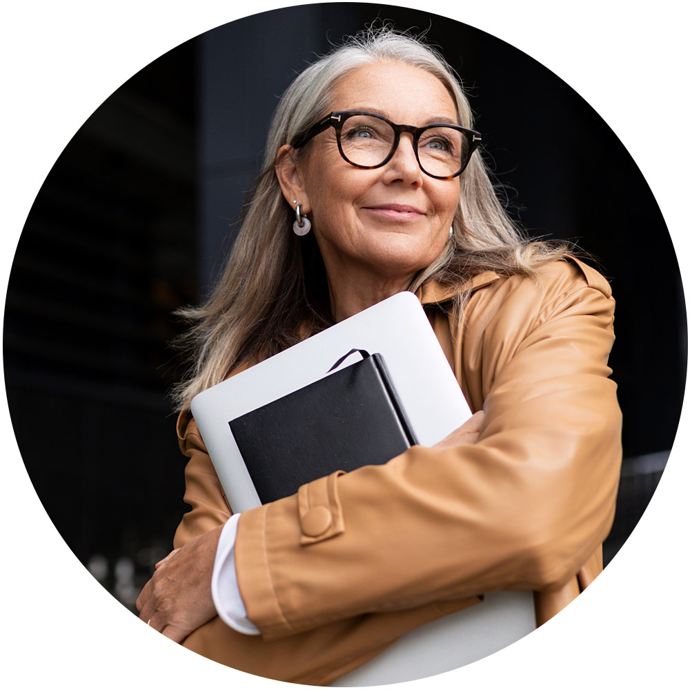 Eine Frau mit langen grauen Haaren und Brille lächelt selbstbewusst und verkörpert Employer Branding. Sie hält einen Laptop und ein Notizbuch in der Hand. Sie trägt einen hellbraunen Mantel und steht gelassen vor einem dunklen Hintergrund.