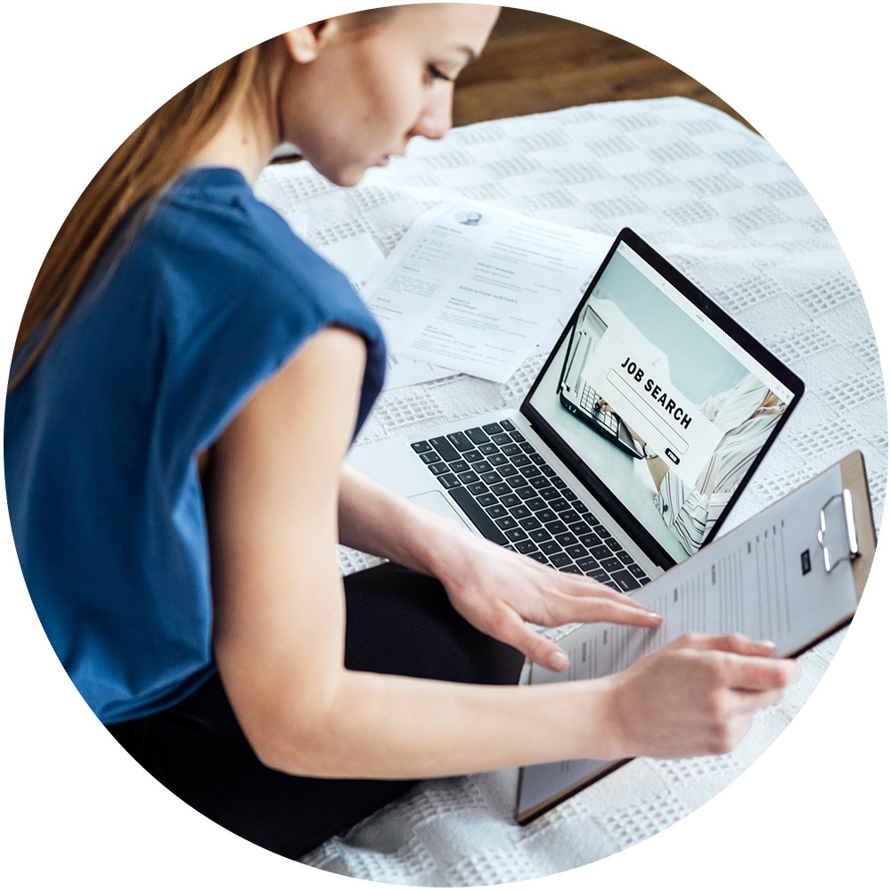 Eine Frau sitzt auf einem Bett und benutzt ihren Laptop mit einer geöffneten Webseite zur „Stellensuche“. Sie verkörpert damit den Geist einer professionellen Personalvermittlerin. Sie hält ein Klemmbrett mit Papieren in der Hand, während die helle Beleuchtung der ungezwungenen Umgebung herrscht.