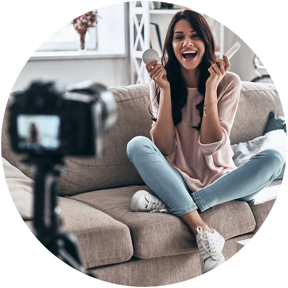 Eine Frau mit langen dunklen Haaren sitzt im Schneidersitz auf einem Sofa, lächelt und hält in jeder Hand Make-up-Artikel. Eine Kamera auf einem Stativ rahmt das Bild perfekt ein und fängt den Moment ein, während sie Videos dreht. Sie trägt einen rosa Pullover, hellblaue Jeans und weiße Turnschuhe.