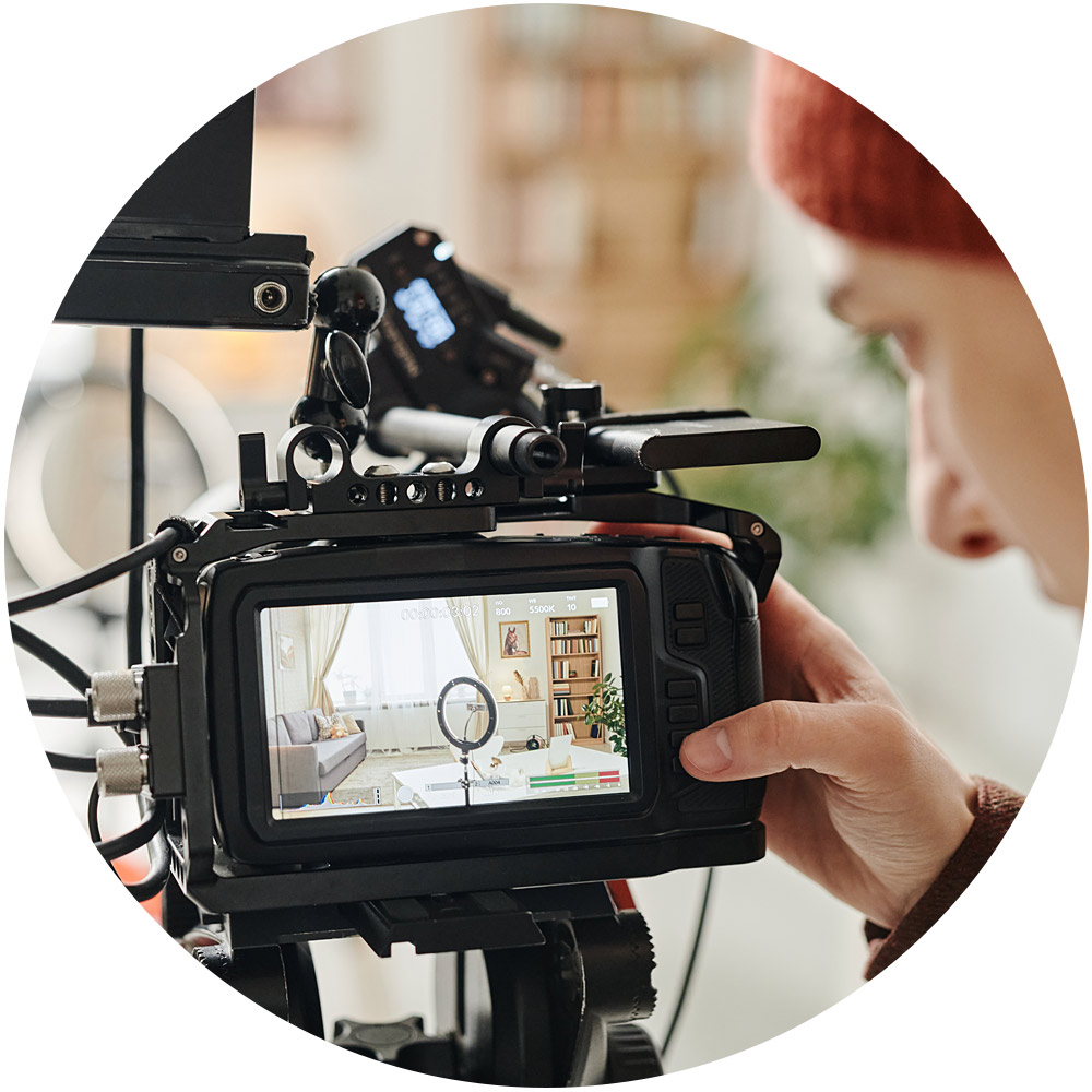 Eine Person bedient gekonnt eine professionelle Videokamera und fängt die Wohnzimmerszene ein, die als scharfes Bild auf dem Bildschirm angezeigt wird. Der Raum ist mit sanftem Licht, einem runden Tisch, einer Pflanze und einem Stativ ausgestattet.