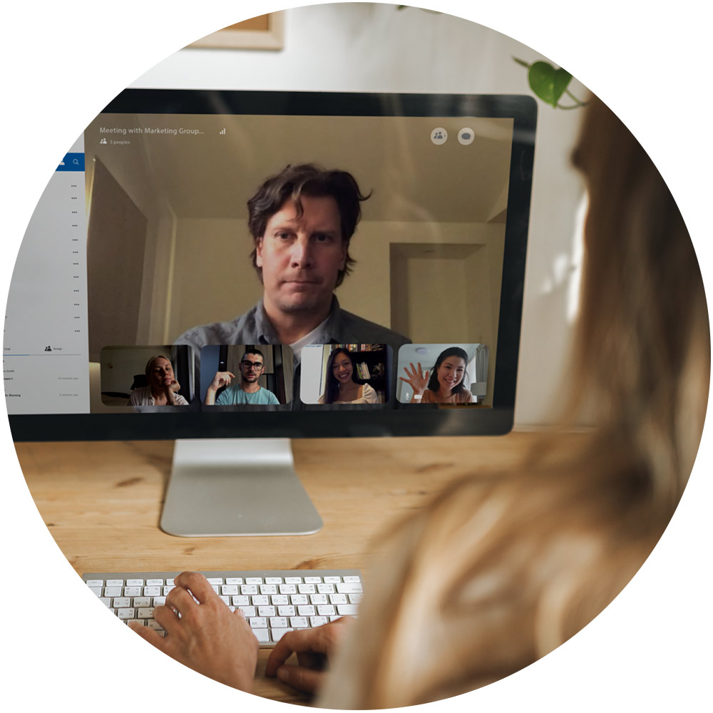 Eine Person nimmt an einer Videokonferenz über einen Computer teil. Auf dem Bildschirm sind mehrere Kollegen zu sehen, die während des virtuellen Meetings lächeln und miteinander interagieren. Auf dem Schreibtisch steht eine Tastatur und in der Nähe sind grüne Pflanzen zu sehen, die dem digitalen Treffen des Newsletter-Teams eine erfrischende Note verleihen.