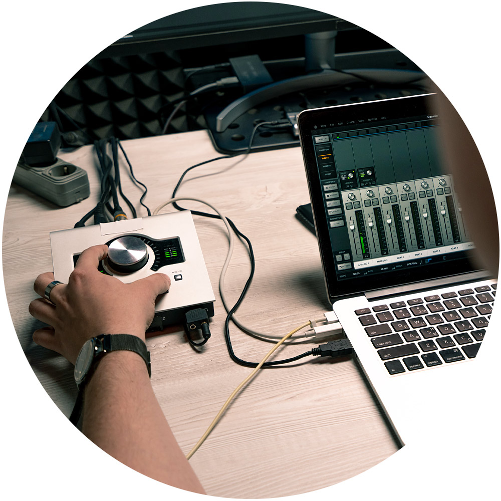 Eine Person justiert einen Regler an einem Audio-Interface, das an einen Laptop angeschlossen ist. Auf dem Bildschirm ist eine Audio-Mixing-Software mit verschiedenen Schiebereglern zu sehen, die eine Technologie zum Klonen von Stimmen nahtlos integriert. Das Setup steht auf einem Holzschreibtisch und ist von einem Kabelgewirr umgeben.