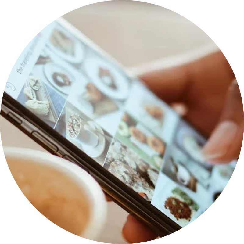 Eine Person hält ein Smartphone in der Hand und durchsucht eine Galerie mit köstlichen Essensfotos. Auf dem Bildschirm werden verschiedene Gerichte angezeigt, während im Vordergrund eine verschwommene, dampfende Tasse Kaffee steht. Es ist eine perfekte Mischung aus Social-Media-Genuss und kulinarischer Kunst, die zum Interagieren und Entdecken einlädt.