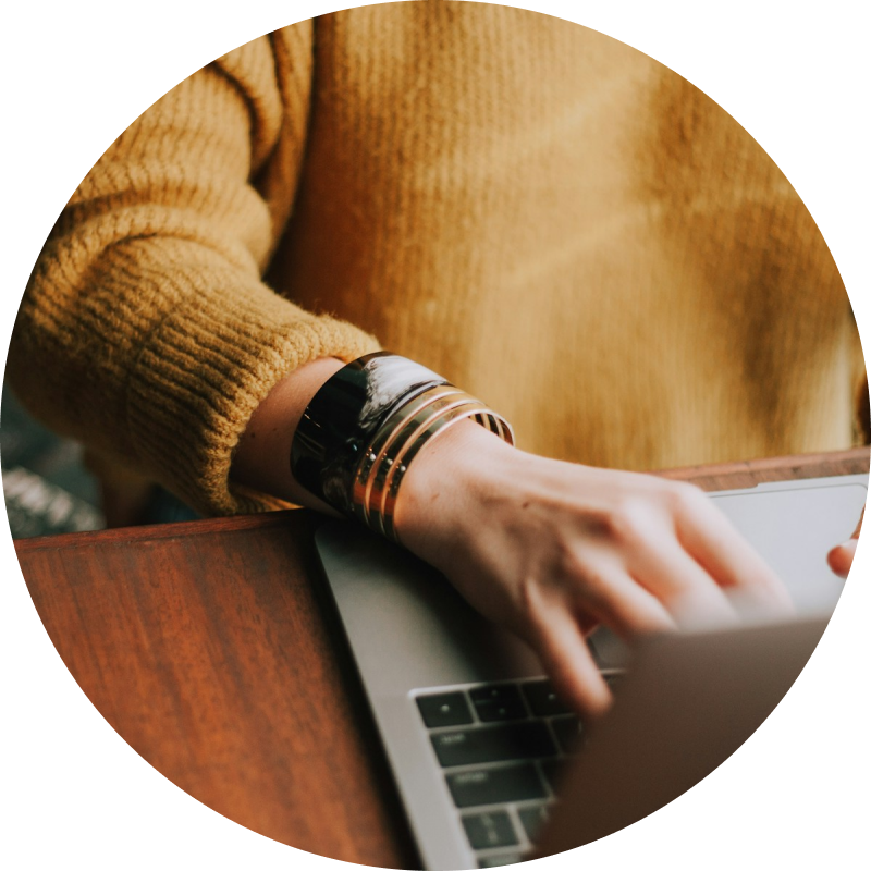 Eine Person in einem senffarbenen Pullover tippt auf einem Laptop und verwaltet möglicherweise soziale Medien. Sie trägt ein großes, glänzendes Armband am Handgelenk. Die Szene ist drinnen, in der Nähe steht eine Holztischfläche.