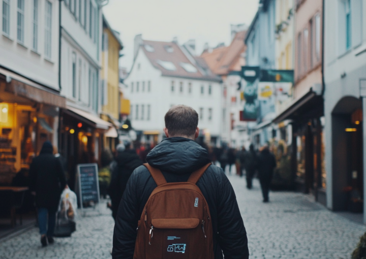 Eine Person mit einem braunen Rucksack spaziert eine Kopfsteinpflasterstraße entlang, die von bunten Geschäften gesäumt ist. Der Himmel ist bewölkt, und das geschäftige Treiben verleiht der lebendigen Stadt einen besonderen Charme, der an eine SEO-Agentur in Ravensburg erinnert. Im Hintergrund schlendern Menschen gemächlich und fügen der Szenerie Lebendigkeit hinzu.