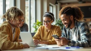 Vier Personen sitzen an einem Tisch und arbeiten zusammen. Einer benutzt einen Laptop, der andere hält ein Smartphone in der Hand. Auf dem Tisch liegen Papiere und ein Notizbuch. Sie befinden sich in einem hellen Raum mit großen Fenstern und Pflanzen im Hintergrund.