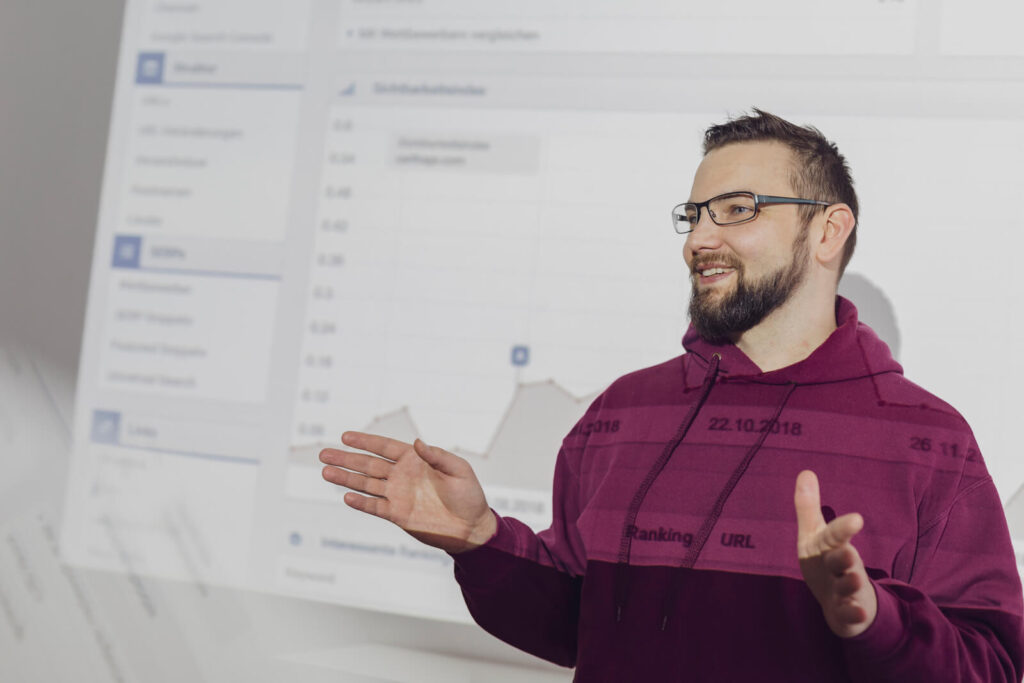 Ein Mann in einem kastanienbraunen Kapuzenpullover und einer Brille gestikuliert, während er vor einer Projektionsfläche spricht, auf der ein Diagramm und Daten angezeigt werden, die die Erfolgskennzahlen seiner WordPress-Agentur veranschaulichen.