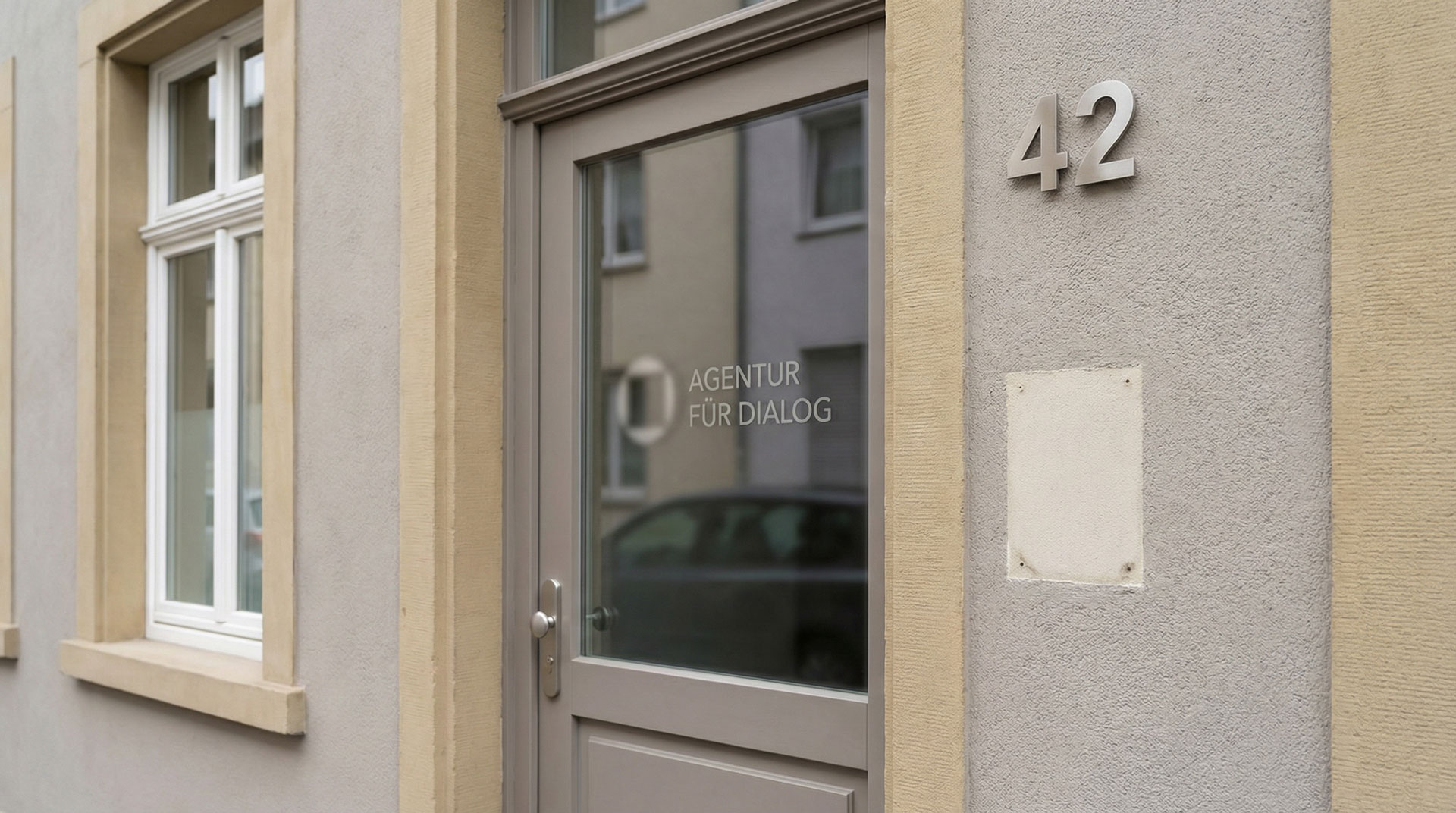 Eine moderne graue Tür mit dem Schriftzug "Agentur für Dialog" auf dem Glas, ideal für B2B-Projekte, steht neben einer beigen Hauswand mit der Nummer 42 und einem Fenster, in dem sich die benachbarten Gebäude spiegeln.